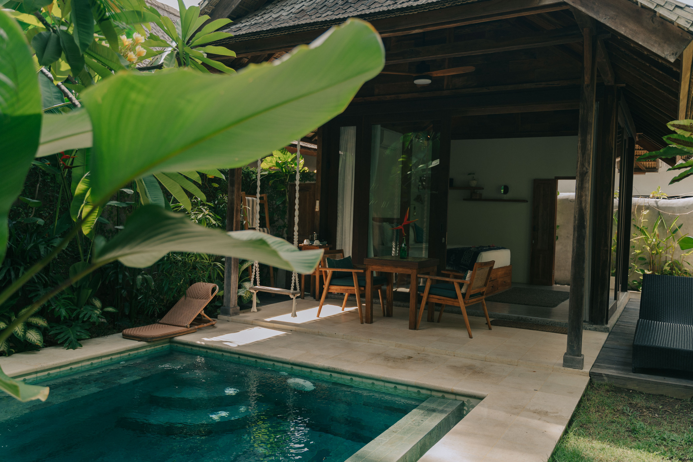 A tranquil villa in Jati Lombok, featuring a private pool surrounded by lush tropical greenery. A charming wooden swing hangs from the pergola, complementing the rustic outdoor seating area with a wooden table and chair. The perfect blend of relaxation and nature.